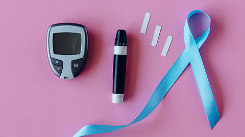 A glucose meter, lancing device, test strips, and light blue ribbon on a pink background.