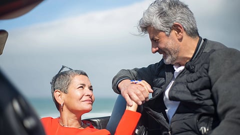 An older man and woman sit together in a convertible by the ocean.