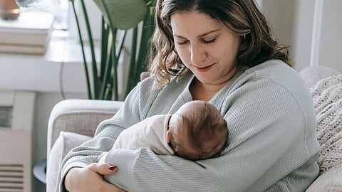 A mother in a sage sweater gently cradles her newborn on a beige couch.