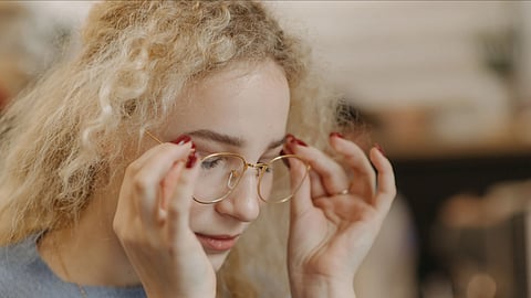 Close-up of a woman with curly hair adjusting her eyeglasses in a warm, blurred indoor setting