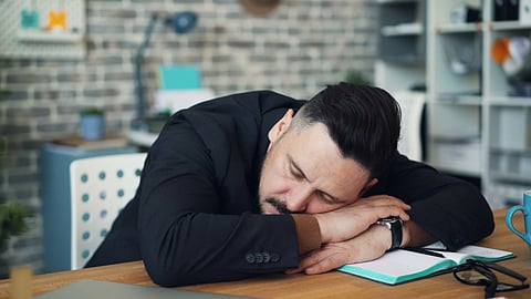 Exhausted man in office resting on desk with notebook and coffee mug