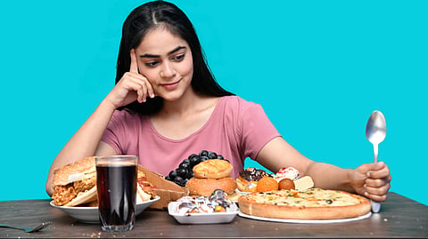  A woman wearing a pink t-shirt holding a spoon looking at a large variety of junk food like burger, pizza, donuts, soft drink.