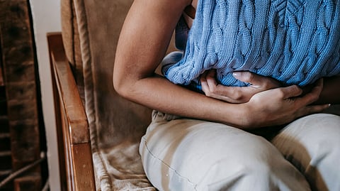 A woman sitting indoors holding a pillow against her stomach, indicating discomfort or pain
