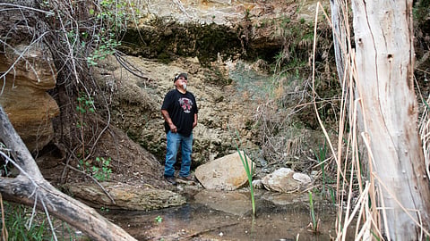 An elderly man in a black T-shirt stands near Entrance Spring in Utah, surrounded by rocks and trees. Elevated radionuclide levels were found there, linked to the White Mesa Mill, where uranium ore and radioactive waste were processed.