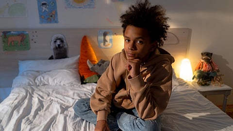 A young boy with curly hair sits on a bed in a cozy bedroom setting, thinkind something
