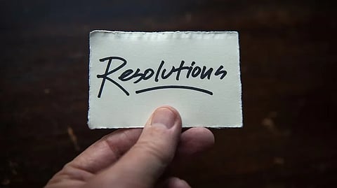 A hand holds a small card with the word "Resolutions" written in bold, black cursive, on a dark, blurred background. 