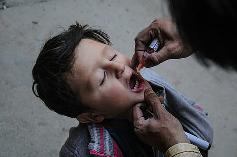 A close-up shot of a young child with eyes closed and mouth open, receiving oral polio vaccine drops from a small plastic vial.