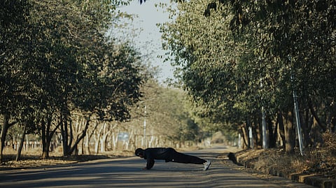 A man performs push-ups outdoors on a tree-lined pathway under the sun