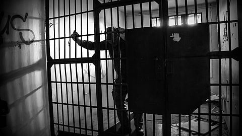 Black and white image of a lone prisoner behind bars inside a dimly lit cell