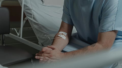 An unrecognizable man in a hospital gown sits on a bed with an IV drip in his arm