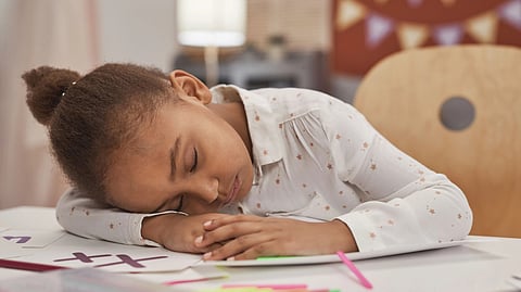 A tired young girl with closed eyes rests her head on homework at a table indoors