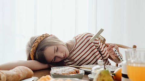 A girl on the food table, looking tired and a fork and piece of food in one hand.