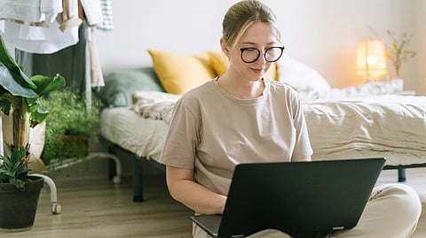 A girl sitting in front of her bed with a laptop on her lap.