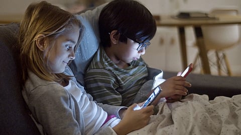 Two children using smartphones at night, indoors, under blankets