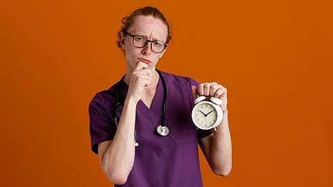 Confused grabbed chin holding alarm clock young male doctor wearing uniform with stethoscope isolated on orange background