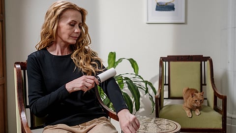 A woman cleaning her clothes with a lint roller while a ginger cat sits nearby, indoors