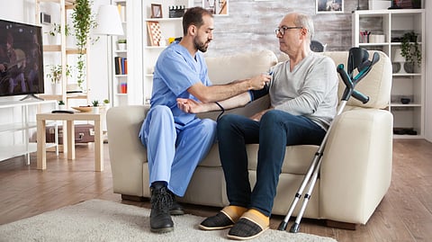 A nurse helping  an old man with the bp machine. Both of them are sat on the sofa.
