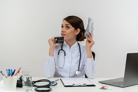 Thoughtful young female doctor wearing medical robe and stethoscope sitting at desk with medical tools and laptop holding credit card and money looking up isolated