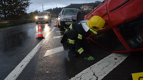 A red car that is turned upside down due to an accident and an emergency worker looking inside the car on the road.