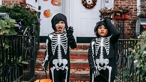 Two kids in skeleton costumes celebrating Halloween with trick-or-treat buckets on a festive porch