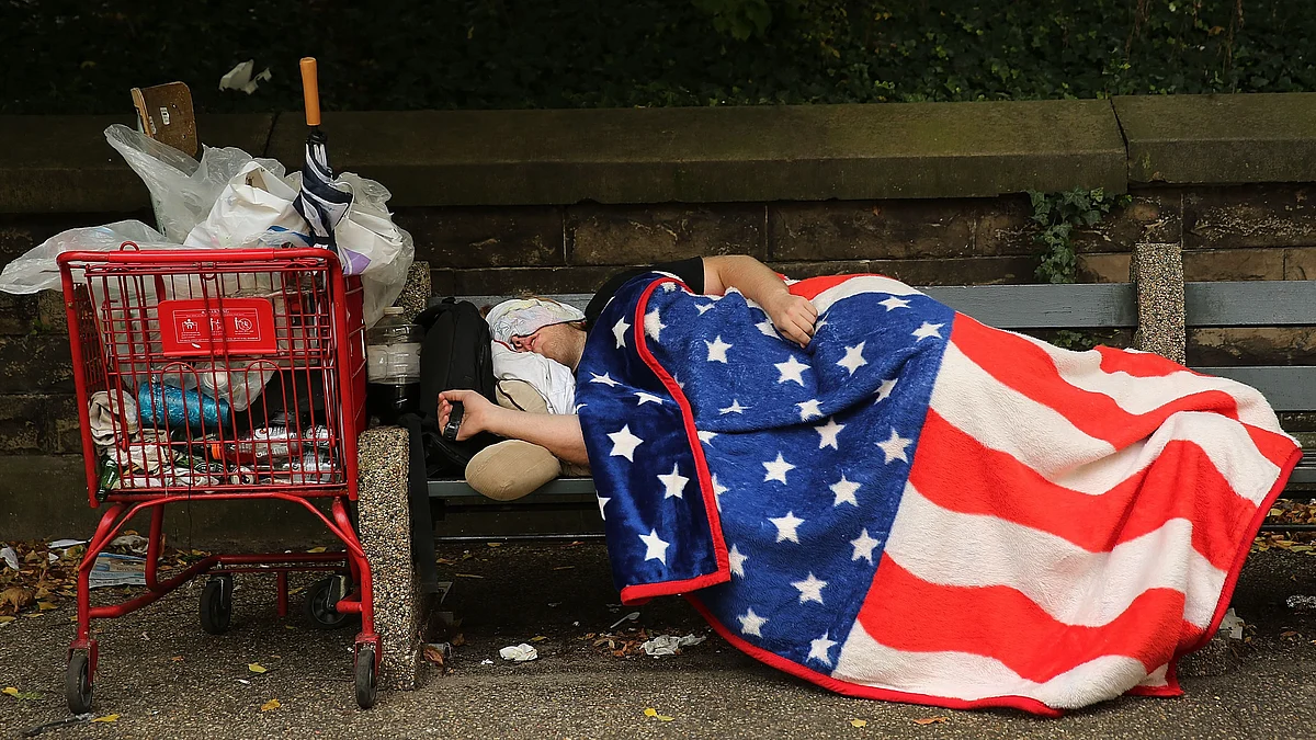 Representative image of a homeless person asleep on a park bench, using a blanket inspired by the US flag. US poverty rate climbs to 12.4%  in 2022, with child poverty more than doubling from 2021 (photo:  Spencer Platt/Getty Images)