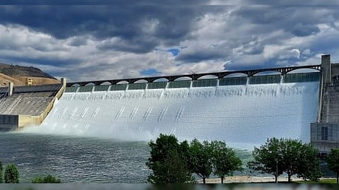 Large dam with water flowing over its spillway under a cloudy blue sky.