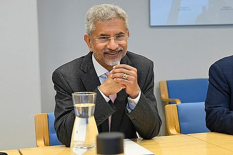 A man in a suit sitting at a table, smiling with his hands clasped, and a glass of water in front of him.