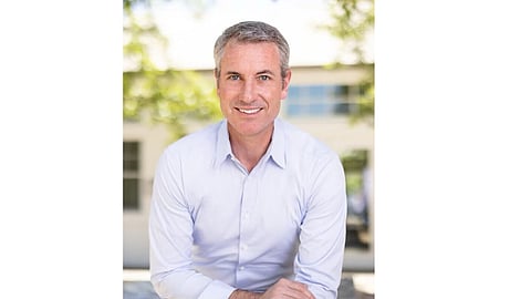 The image shows a man in a light-colored shirt smiling while sitting outdoors.