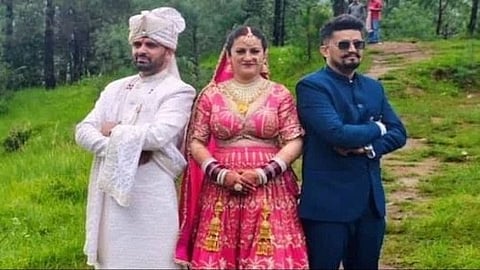 The image shows a bride in a red and pink lehenga standing between two men dressed in traditional and formal attire, posing outdoors.