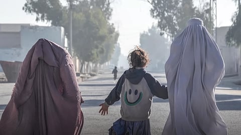 Three women walking together down a city street, surrounded by urban buildings and a clear blue sky.
