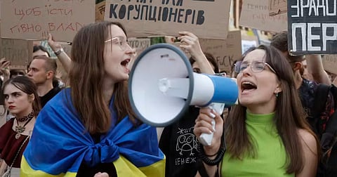 Woman shouting into a megaphone during a protest, draped in a blue and yellow flag.