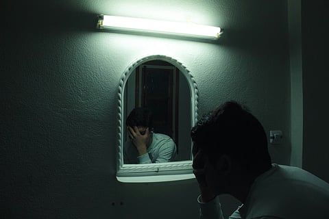 A person’s reflection appears in a small mirror under dim light with a fluorescent lamp above.
