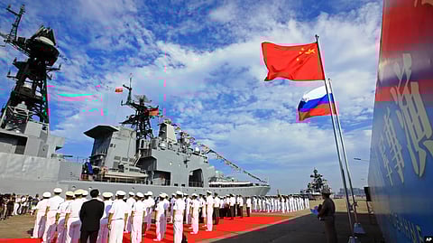 In the image officers are shown standing in front of a ship and the flag of Russia and China are hoisted