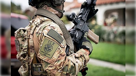 A person in camouflage gear with an ICE badge and patch, holding a rifle, stands outdoors in front of a building and greenery.


