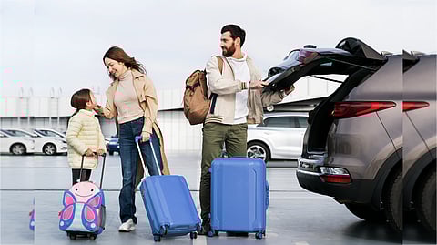 A family prepares for travel at a parking lot. A man loads blue suitcases into a car trunk, while a woman and child with a butterfly suitcase smile nearby.