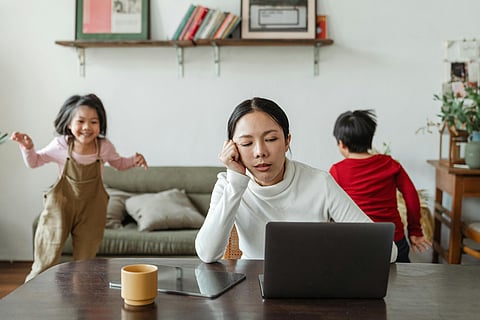 An image of a living room in which a woman is working in her laptop and the kids are playing in the background