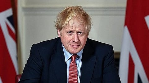 Boris Johnson sitting in his office wearing blue coat and behind him in both sides are the flags of Britain
