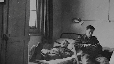 In the image two soldiers are shown in their room. Both are in their uniform one is reading book while the other is sleeping in a small bed