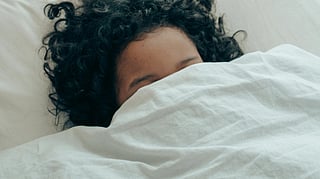 A girl in her bed. The bed is white and she is hiding her face with her sheet