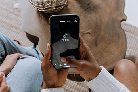 Image of a woman holding to her phone which has the tik tok open on the screen. In the background there is a wooden table.