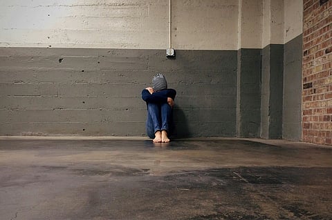 In the image a girl is shown in an empty room with her head in her knees 