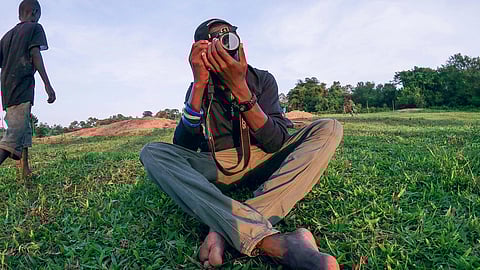 In the image someone is shown sitting in green field with a camera in their hands and another person is walking from the back
