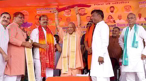 In the image yogi Adityanath is shown raising his hands and there are other leaders present in stage addressing audience