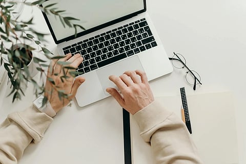 Image of a man typing on his laptop with his spectacles, coffee and notepad on his table.