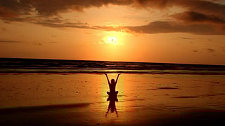 In the image a person is shown in a beach with hands raised the time is of evening with sunset background