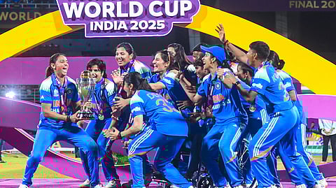 Indian Women's Cricket team celebrating their historic win with the ICC World Cup trophy. They are all happy with the World Cup India 2025 logo in the background.