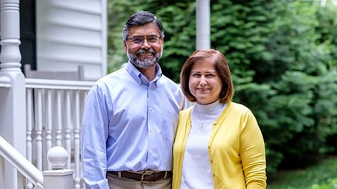 Ghazala Hashmi posing for a picture with her husband, Azhar. She is wearing a white top and a yellow cardigan whereas her husband is wearing a blue shirt. Trees and a porch are visible in the background. 