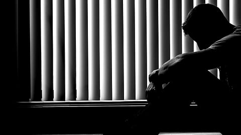 Silhouette of a man seated on the floor by vertical blinds, head bowed and arms on knees. The dim lighting and posture convey a somber, introspective mood.
