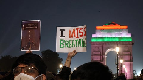 Protesters in masks hold signs reading "I Miss Breathing" and "Tintin in Delhi" at night. India Gate is lit in flags' colors, conveying urgency.