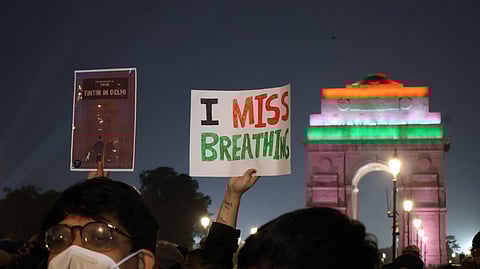 Protesters in masks hold signs reading "I Miss Breathing" and "Tintin in Delhi" at night. India Gate is lit in flags' colors, conveying urgency.
